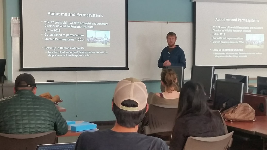 A man named Chris Meador standing in front of a group of students sitting in chairsand giving a presentation about permaculture with two powerpoint slides being projected on either side of him in San Diego, California