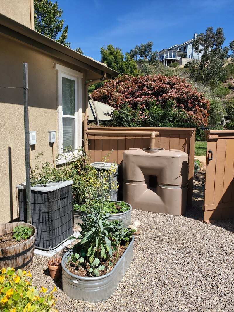 Side yard of house with tan 265 gallon Bushman water tank connected to roof with pipes to collect rainwater and distribute it to small vegetable garden in metal troughs located in Scripps Ranch, San Diego