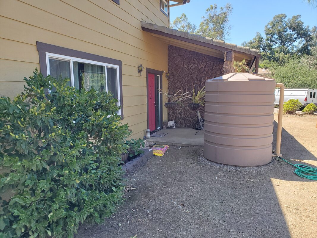 Tan 865 gallon Bushman rainwater tank connected to house gutters to catch water and a garden hose to distribute water from the tank