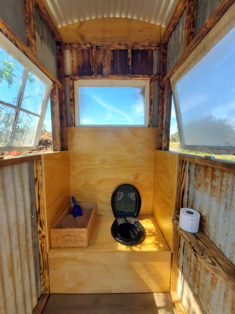 Inside of the compost toliet building, bog windows bring in lots of light and ventilation with a black toliet seat and a box of sawdust is used to cover deposits
