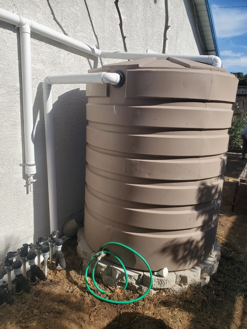 865 gallon mocha Bushman water tank elevated on blocks connected to home gutters with pipe, first flush device and garden hose to ditibute water captured in tank